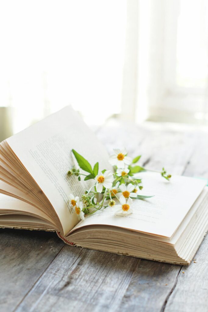 A serene image of an open book with delicate wildflowers on a wooden table by a sunlit window.